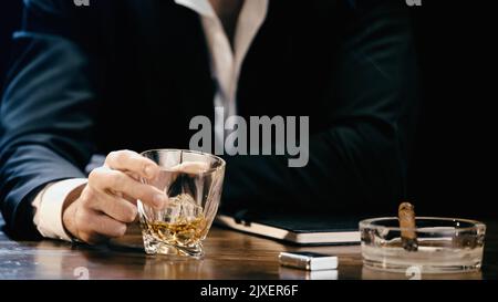 Beschnittene Ansicht des Geschäftsmannes, der ein Glas Whiskey in der Nähe einer Zigarre in einem Aschenbecher hält, isoliert auf schwarzem, stockem Bild Stockfoto