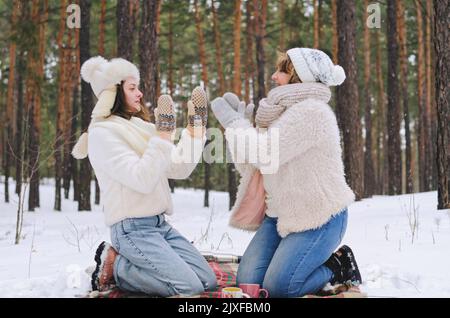 Familie in verschneiten Park, Wald. Junge Frau, Mädchen und ihre Mutter sitzen in der Nähe von schneebedeckten Kiefern. Picknick. Kaffee trinken. Spaß haben. Stockfoto