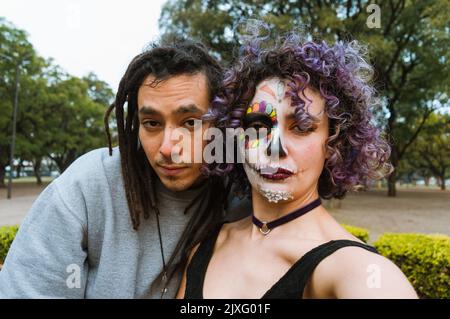 Porträt eines jungen lateiners und einer jungen Frau im Freien, die einen Tag des toten Selfies mit einem bemalten catrina-Gesicht in einem Park mit einem Lächeln und Blick auf die Kamera machen Stockfoto