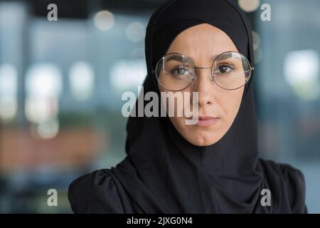 Nahaufnahme einer Geschäftsfrau in schwarzem Hijab, einer muslimischen Frau, die bei der Arbeit im modernen Bürogebäude arbeitet und mit Konzentration und ernster Nachdenklichkeit in der Brille auf die Kamera schaut. Stockfoto