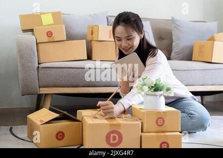 Junge lächelnde schöne Besitzer asiatische Frau Freiberufler kmu Geschäft Online-Shopping Arbeit auf Laptop-Computer mit Paketkasten auf dem Bett zu Hause - KMU Stockfoto