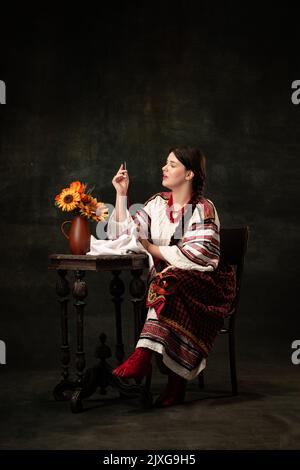 Hübsche junge Frau in volkstümlicher ukrainischer Kleidung, die isoliert auf dunklem Vintage-Hintergrund sitzt. Mode, Schönheit, Kulturerbe, Epochen Stockfoto