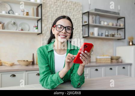 Online-Shopping. Zufriedene schöne junge Frau macht Online-Einkäufe, bestellt nach Hause liefern vom roten Telefon. Er sitzt in der Küche zu Hause in Gläsern und grünen Kleidern und lächelt. Stockfoto