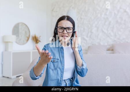 Webinar. Junge, schöne Geschäftsfrau mit Brille und Kopfhörer sitzt zu Hause auf dem Sofa und führt ein Webinar, Online-Schulungen vor der Kamera, berät, erzählt, Gesten mit den Händen, lächelt. Stockfoto