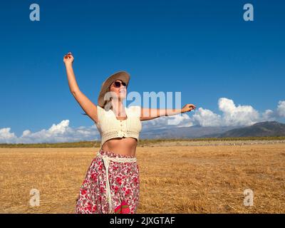 Positive Frau in böhmischer Kleidung mit Hut und Sonnenbrille, die die Hand hebt und den Arm ausstreckt, während sie auf dem trockenen Feld gegen den wolkigen blauen Himmel steht Stockfoto