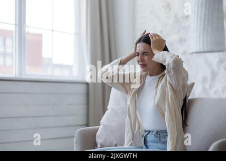 Nervenzusammenbruch, Depression, Panikattacke. Eine junge, schöne Brünette sitzt zu Hause auf dem Sofa und hält ihren Kopf mit den Händen, ist nervös, fühlt Schmerzen, Beschwerden. Stockfoto