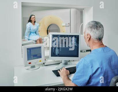 Radiologe vor dem CT-Scan des weiblichen Körpers mit CT-Scanner aus dem Kontrollraum hinter dem Schutzfenster. Computertomographie Stockfoto