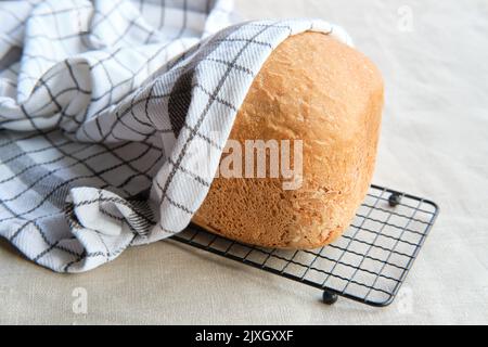 Hausgemachtes Bauernhaus gemischtes Brot. Nahaufnahme von Brot, das in der Backmaschine gebacken wurde. Vollkornbrot auf Trockengestell mit gestreifem Handtuch auf Textil Stockfoto