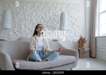 Zeit für Yoga. Attraktive junge Frau mit Brille sitzt auf dem Sofa zu Hause in Lotusposition mit geschlossenen Augen. Ruht sich aus, entspannt sich. Stockfoto