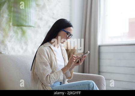 Bloggen. Freiberufler. Zufriedene junge schöne Frau in Brille sitzt zu Hause auf dem Sofa am Fenster und nutzt das Telefon, Typen, schreibt einen Beitrag, Blogs. Stockfoto