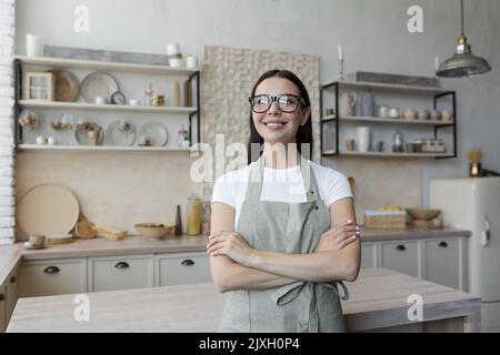 Porträt einer jungen schönen Frau Köchin, Konditorin. Sie steht in einer beigefarbenen Schürze und Brille in der Küche, ihre Arme gekreuzt. Er schaut auf die Kamera und lächelt. Stockfoto