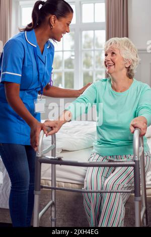Ältere Frau Zu Hause, Die Den Gehrahmen Benutzte, Wurde Von Einer Weiblichen Pflegekraft In Uniform Aus Dem Bett Geholfen Stockfoto