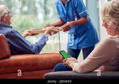 Nahaufnahme Einer Älteren Frau, Die Das Mobiltelefon Nutzt, Während Die Betreuerin In Der Lounge At Home Eine Mahlzeit Zum Senior Man Bringt Stockfoto