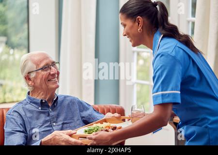 Weibliche Pflegekraft In Uniform, Die Essen Auf Tablett Zum Älteren Mann Bringt, Der In Der Lounge Zu Hause Sitzt Stockfoto