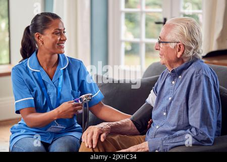 Älterer Mann Zu Hause In Der Lounge, Der Von Einer Weiblichen Pflegekraft In Uniform Einen Blutdruck Genommen Hat Stockfoto