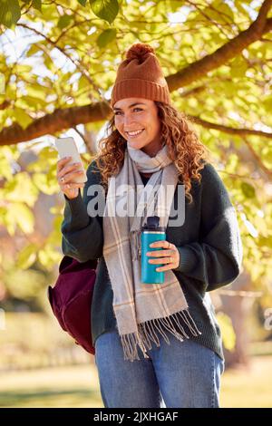 Lächelnde Junge Frau Mit Hut Und Schal Im Autumn Park Mit Mobiltelefon Stockfoto