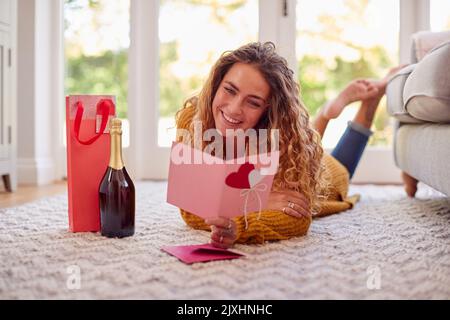Frau im warmen Pullover, die zu Hause auf dem Boden liegt und die Valentinskarte mit einer Flasche Champagner liest Stockfoto