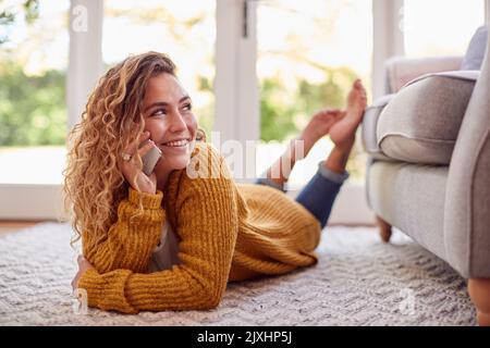 Frau Im Warmen Pullover, Die Zu Hause Auf Dem Boden Liegt Und Auf Dem Mobiltelefon Spricht Stockfoto