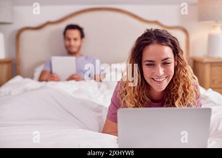 Paar Tragen Pyjamas Liegen Im Bett Zu Hause Mit Frau Mit Laptop Und Mann Digital Tablet Stockfoto