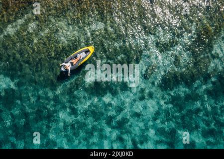 Ein einsames Weibchen in einem Strohhut lächelnd, entspannt im Kajak auf den türkisfarbenen Wellen des Adriatischen Meeres liegend. Luftaufnahme der Küste von oben. Exotisches c Stockfoto