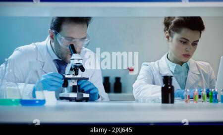 Wissenschaftler in einer Brille, die im Labor durch das Mikroskop schaut, in der Nähe eines Kollegen, der einen Laptop benutzt, Stockbild Stockfoto