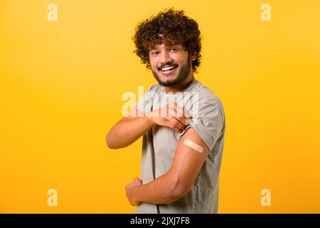 Lächelnder junger lockiger Inder, der nach der Impfstoffinjektion auf gelbem Hintergrund einen Arm mit Bandenhilfe zeigt, fühlt sich der geimpfte multirassische Mann mit dem Impfstoff sicher Stockfoto