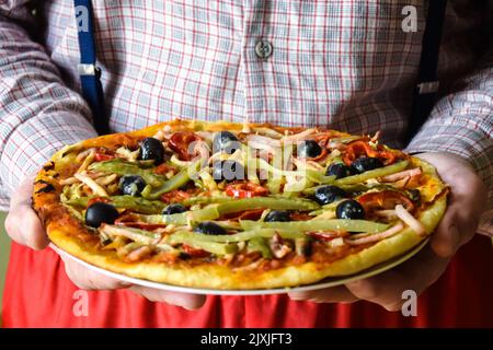 Ein Mann hält frisch zubereitete Pizza in den Händen. Hausgemachte italienische hausgemachte Pizza: Oliven, Schinken, Pfeffer, Sauce, Gurke, Käse. Nahaufnahme. Se Stockfoto