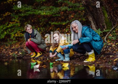 Kleines Mädchen mit Mutter und Großmutter, die mit Papierbooten in See-Outoors in der Natur spielen. Stockfoto