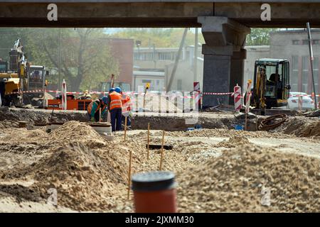 RIGA, LETTLAND - 13. MAI 2022: Eine Gruppe von Arbeitern in der Uniform stand auf der Baustelle in einem gegraben Loch während des neuen Straßenbaus unter der Brücke. Stockfoto