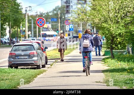 RIGA, LETTLAND - 13. MAI 2022: Frau, die auf einem Bürgersteig Fahrrad fährt. Ein Mann mit Mantel und Sonnenbrille geht an einem sonnigen Tag in die entgegengesetzte Richtung. Stockfoto