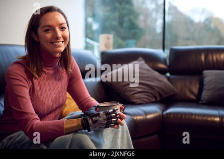 Porträt Einer Frau Mit Prothetischem Arm Und Hand, Die Sich Zu Hause Auf Dem Sofa Entspannt Und Eine Tasse Kaffee Trinkt Stockfoto