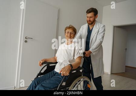 Professioneller männlicher Arzt, der weibliche Patientin auf dem Rollstuhl in der Medizinklinikhalle trägt Stockfoto