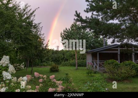 Kleines Landhaus an einem Sommerabend. Regenbogen über dem Haus. Wunderschöner Sonnenuntergang. Hortensien Blumen im Garten Stockfoto