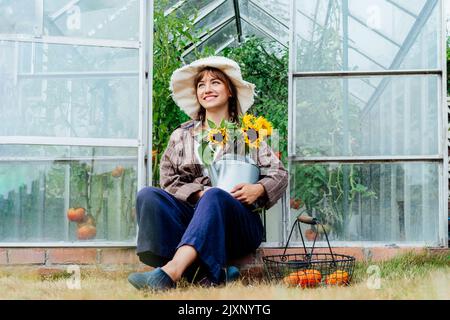 Lächelnde junge Bäuerin hält Gießkannen mit frischem Sonnenblumenstrauß, sitzt in der Nähe von Gewächshaus mit gerade gepflückten Tomaten im Korb. Cottagecore Stockfoto