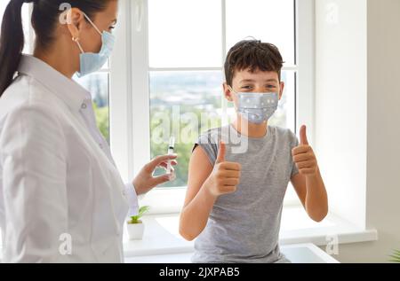 Tenn-Junge zeigt Daumen nach oben vor der Impfung in der Klinik und schaut auf die Kamera, die in der Nähe des Arztes sitzt. Stockfoto