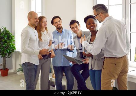 Ein freundlicher männlicher Geschäftsleiter oder Trainer spricht mit einer Gruppe von Personen, die in seiner Nähe im Büro stehen Stockfoto