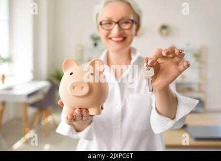 Eine glückliche Frau, die Geld gesparten hat, hält ein Sparschwein und Schlüssel zu ihrem neuen Haus Stockfoto