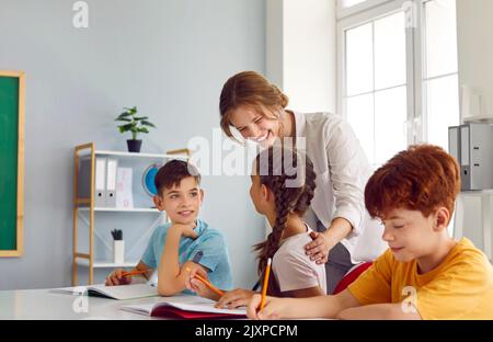 Freundlich lächelnde Lehrerin, die ihren kleinen Schülern während des Unterrichts im Klassenzimmer hilft. Stockfoto