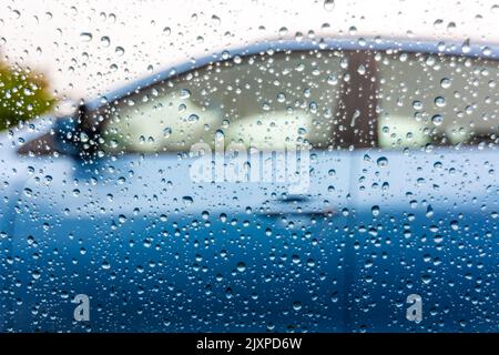 Wasser tropft auf das Autofenster. Blick vom Auto mit Regentropfen am Fenster zum nächsten Auto mit blauer Farbe. Regentag. Stockfoto