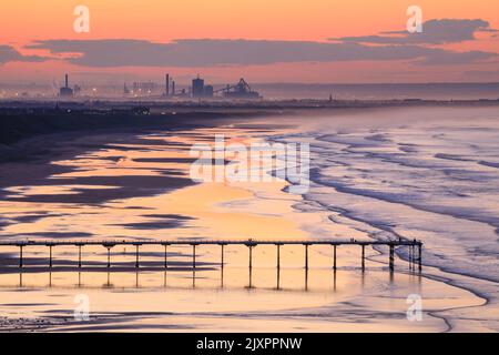 Der Saltburn Pier wurde kurz vor Sonnenuntergang vom Küstenpfad in der Nähe von Saltburn-by-the-Sea mit dem Stahlwerk in Redcar in der Ferne aufgenommen. Stockfoto