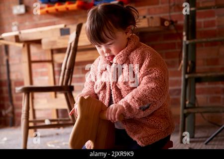 Mädchen Mit Down-Syndrom Sitzen Und Spielen Auf Hölzernen Schaukelpferd In Garage Workshop Zu Hause Stockfoto