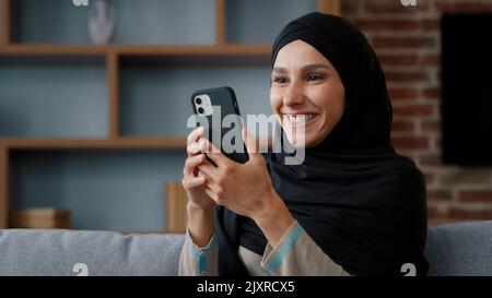 Lächelnd arabisch muslimisch islamisch 30s Frau Benutzer im Kopftuch schwarz Hijab mit Telefon zu Hause sitzen auf Couch Middle Eastern Mädchen Dame mit Handy Stockfoto