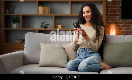 Lächelnd Hispanic Caucasian 30s Frau Hausfrau Mädchen sitzt auf Couch gemütliches Sofa zu Hause mit Smartphone für die Bestellung Lieferung von Lebensmitteln chatten mit Stockfoto