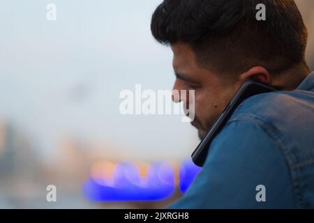 Der arabische Mann hält das Mobiltelefon an Ort und Stelle, indem er es zwischen die linke Schulter und das linke Ohr drückt. Nahaufnahme eines erwachsenen Mannes mit Freisprechfunktion und Kopfhörern. Stockfoto