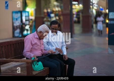 Zwei alte Männer, die eine Brille, Knopfhemden und Slacks tragen, sitzen auf einer Bank und inspizieren einen Gürtel, den sie gerade aus dem Textilmarkt (arabischer Markt) gekauft haben. Stockfoto