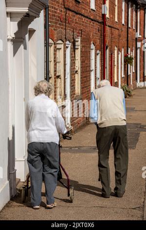 Ein älteres Paar, das am Straßenrand auf einem Gehweg entlang ging, ein altes Paar, das zusammen die Straße entlang ging, eine Dame mit Zimmer-Rahmen und ein Mann, der ging. Stockfoto