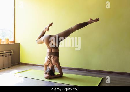 Die junge Frau, die Yoga praktiziert, führt eine Variante der Shirshasana-Übung mit einem gebogenen Bein, einem umgedrehten Asana, durch, steht auf ihren Unterarmen im Studio ne Stockfoto