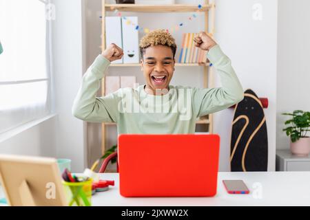 Aufgeregter junger lateinamerikanischer Mann, der gute Nachrichten mit einem Laptop zu Hause feiert Stockfoto