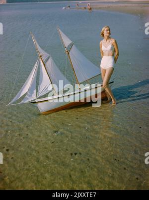 Porträt einer Frau im zweiteiligen Badeanzug, der am Ufer in der Nähe eines Miniatursegelbootes steht, Toni Frisell Collection, 1948 Stockfoto