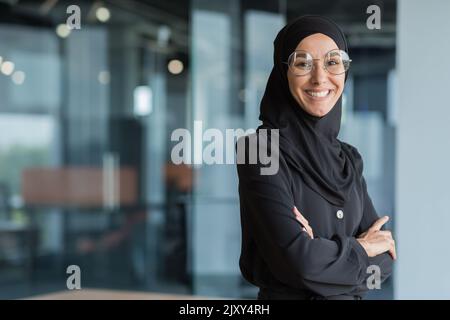 Porträt einer muslimischen Frau in Hijab bei der Arbeit im Büro, einer Geschäftsfrau, die lächelt und mit gekreuzten Armen auf die Kamera schaut. Stockfoto